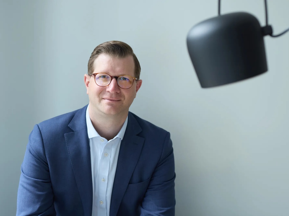 Ein Mann im blauen Anzug sitzt vor einer grauen Wand, eine schwarze Lampe hängt rechts.