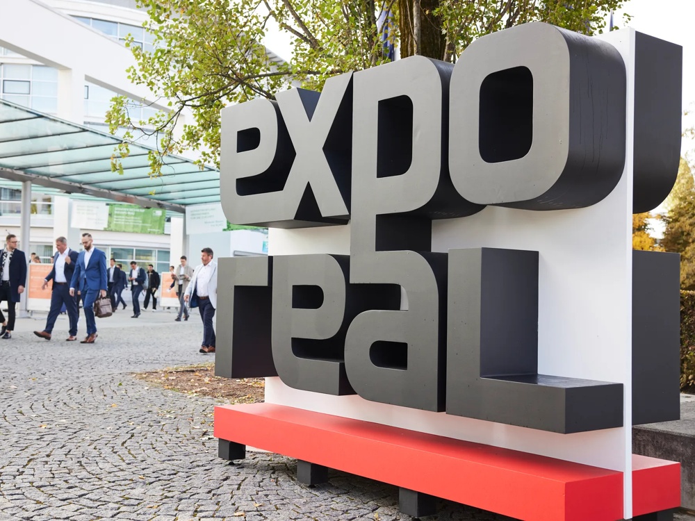 Großes "Expo Real"-Schild im Freien, Menschen in Anzügen gehen auf einem gepflasterten Weg vorbei.