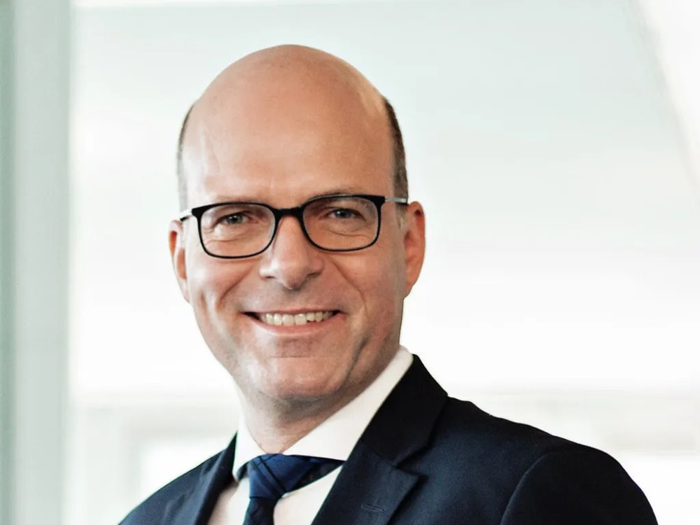 Ein Mann in Anzug und Krawatte lächelt, trägt eine Brille, vor einem hellen Hintergrund.