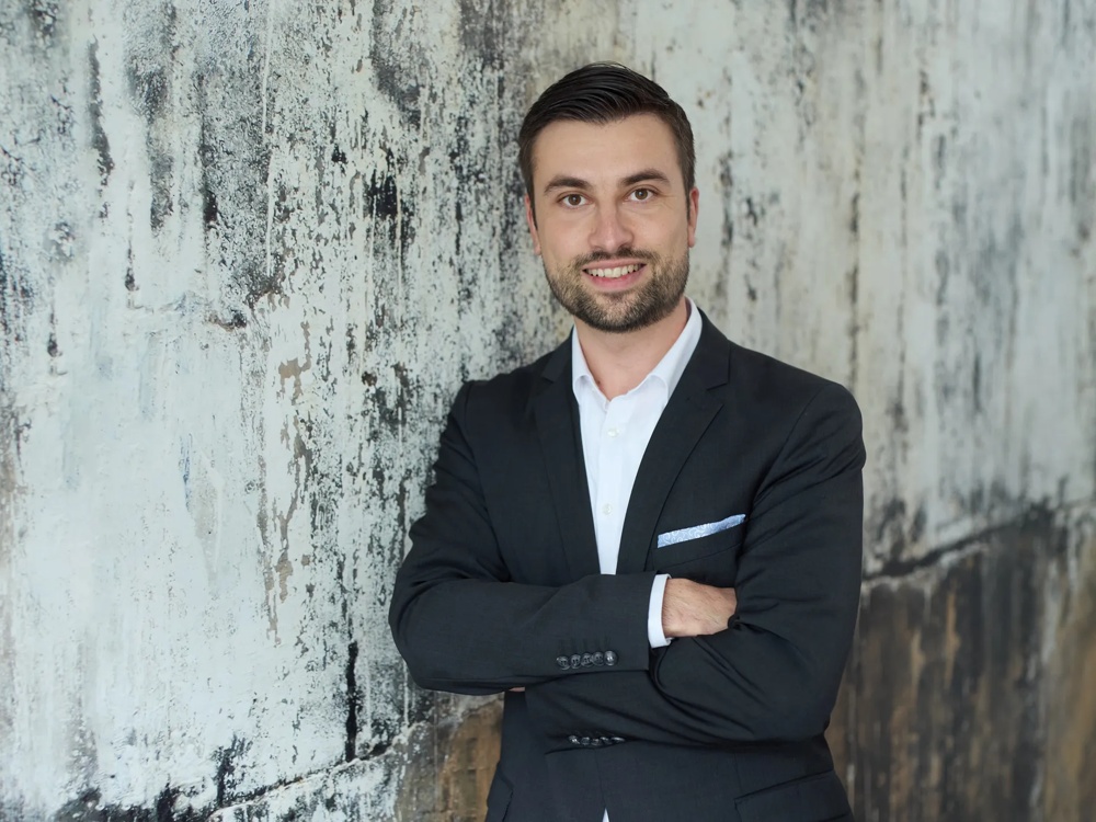 Commerz Real employee Filip Orec, Key Account Manager for Sales Private and Business Clients NOM, is shown. He stands with his arms crossed in front of a textured wall, wearing a dark suit with a white shirt and a light blue hankie.