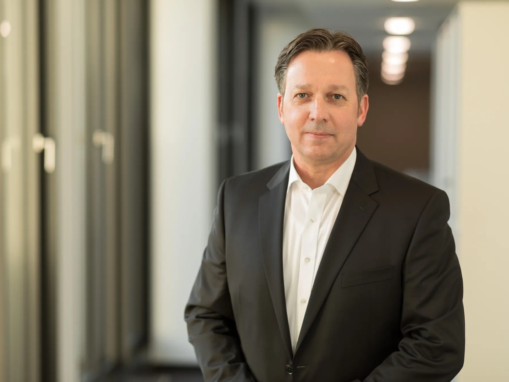 A man in a black suit standing in an office hallway with a blurred background.