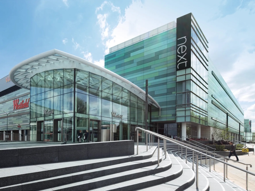 Modernes Einkaufszentrum mit Glasfassade und grünem Gebäude, Treppen im Vordergrund, blauer Himmel im Hintergrund.