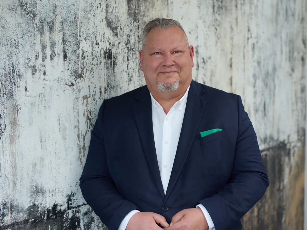 Ein Mann im Anzug steht vor einer verwitterten Wand, trägt ein weißes Hemd und ein grünes Einstecktuch.