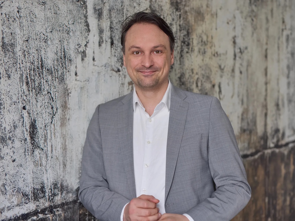 Ein Mann im grauen Anzug steht vor einer strukturierten, verwitterten Wand und lächelt in die Kamera.