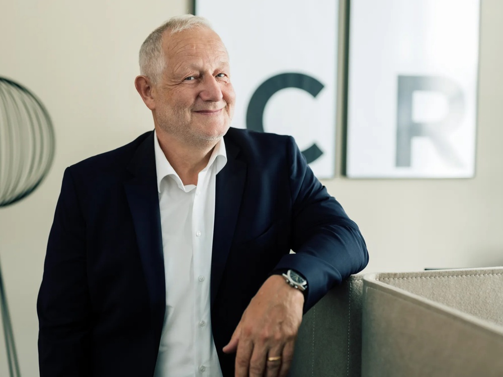 Ein lächelnder Mann im Anzug lehnt an einer Trennwand, im Hintergrund eine Stehlampe und zwei gerahmte Buchstaben.