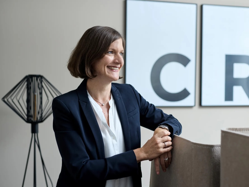 Nadia Eichelberger ist Global Head of Real Estate Office and Industrial. Nadia Eichelberger sitzt vor einem hellen Hintergrund, hinter ihr die gerahmten Initialen der Commerz Real und eine schwarze Stehlampe.