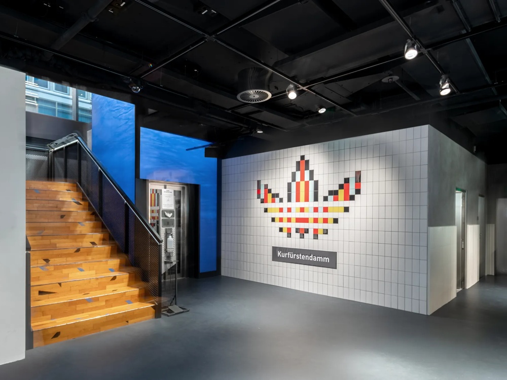 Holztreppe neben weißer Kachelwand mit pixelartigem Logo und "Kurfürstendamm"-Schild, moderner Innenraum.