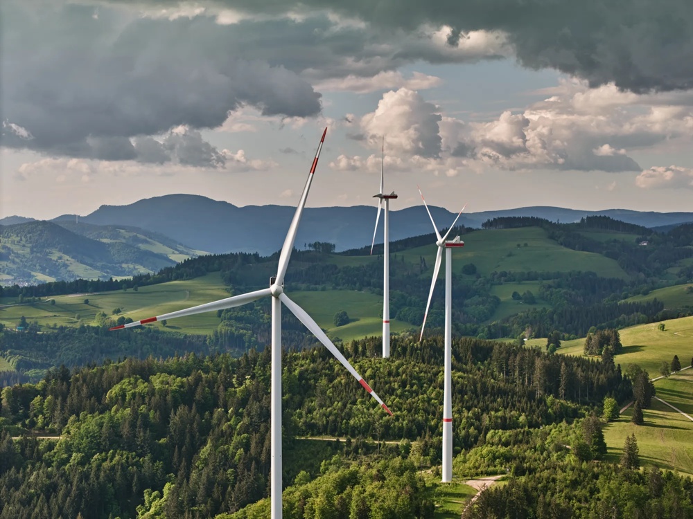 Drei Windräder auf einem bewaldeten Hügel unter bewölktem Himmel, umgeben von grünen Feldern und Bergen.