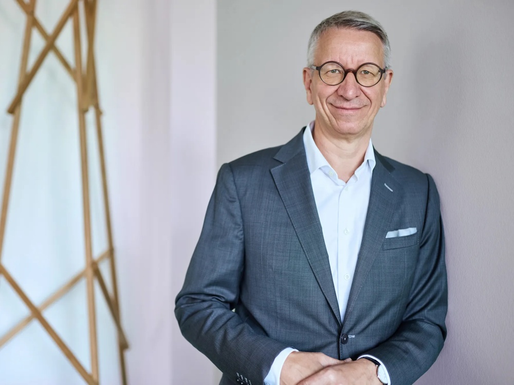 A man in a suit wearing glasses leans against a wall, with wooden struts in the background.