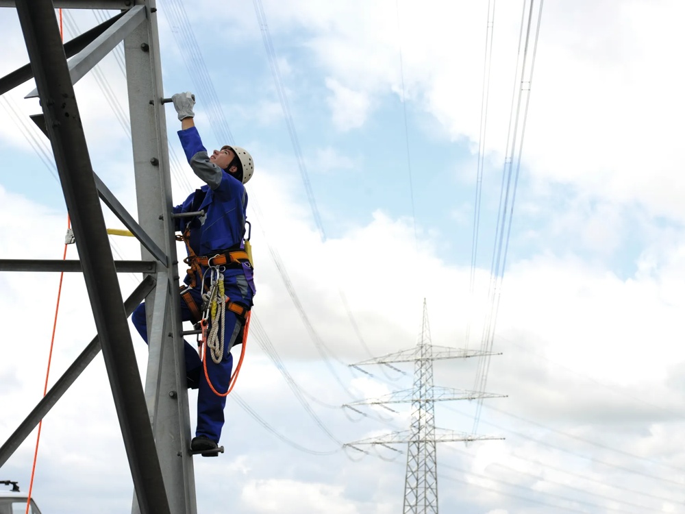 Ein Arbeiter in blauer Schutzkleidung klettert an einem Strommast vor bewölktem Himmel.