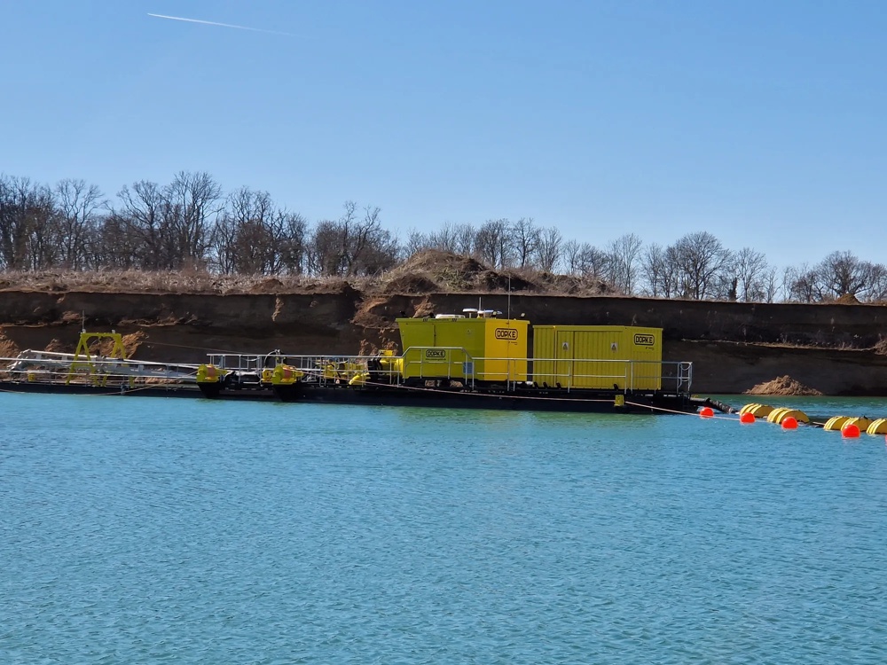 Gelbes Industriegebäude auf einem Floß im blauen Wasser, mit kahlen Bäumen im Hintergrund.