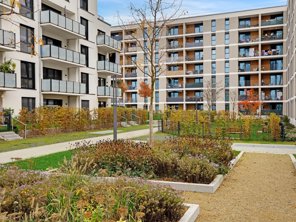 Moderne Wohngebäude umgeben von gepflegten Gärten mit Bäumen und bunten Pflanzen unter blauem Himmel.