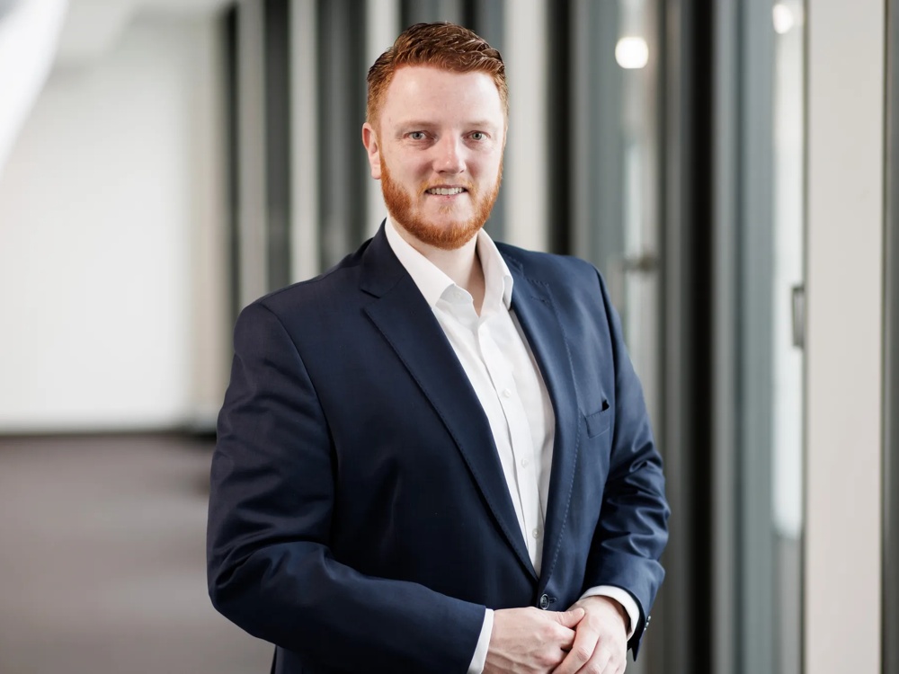 A man in a suit standing smiling in a modern office hallway with large windows.