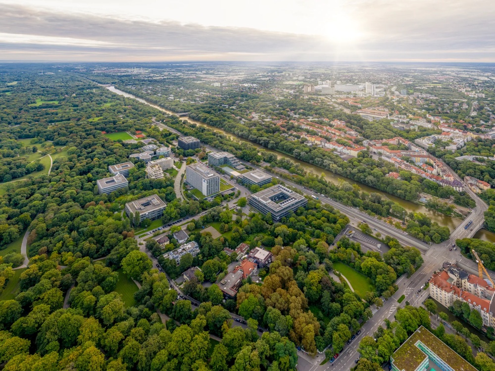 Luftaufnahme eines weitläufigen Parks mit vielen Bäumen, Gebäuden und einem Fluss unter bewölktem Himmel.