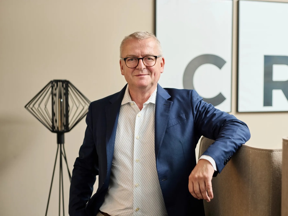 Ein Mann in Anzug steht lächelnd neben einem Sofa, im Hintergrund eine Lampe und zwei Buchstaben-Bilder.