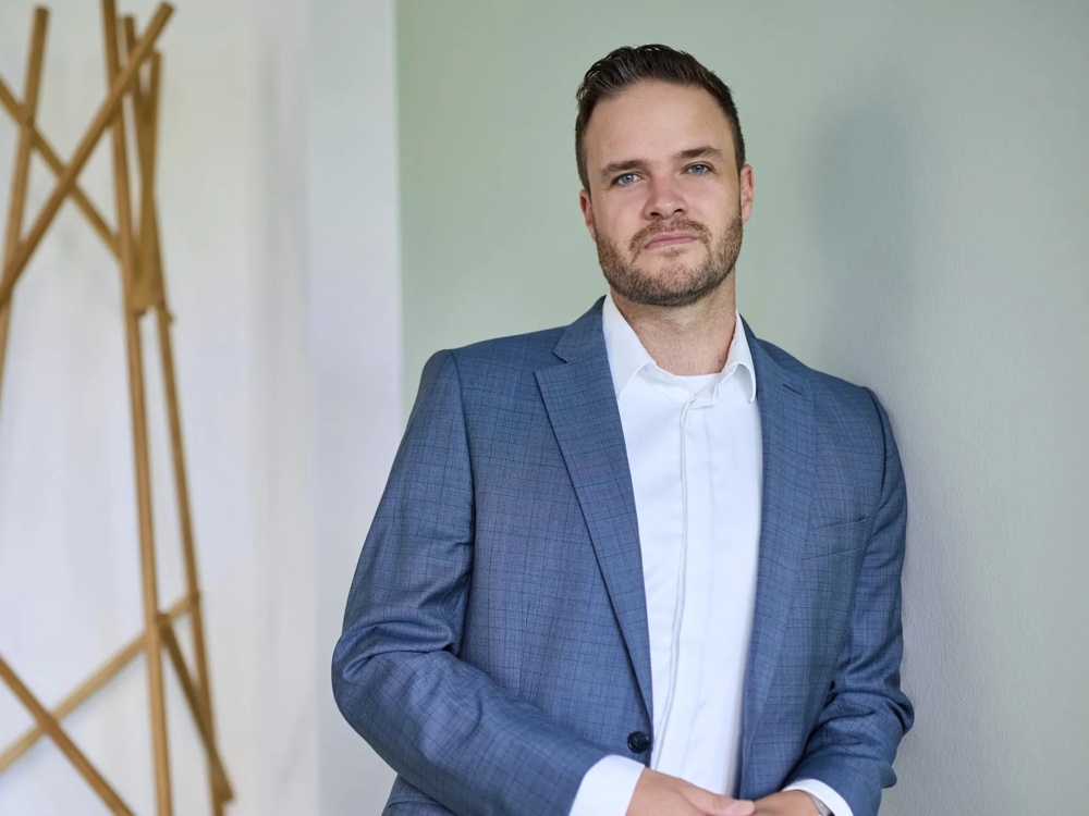 Ein Mann im blauen Anzug steht vor einer weißen Wand, ein Holzkleiderständer im Hintergrund.