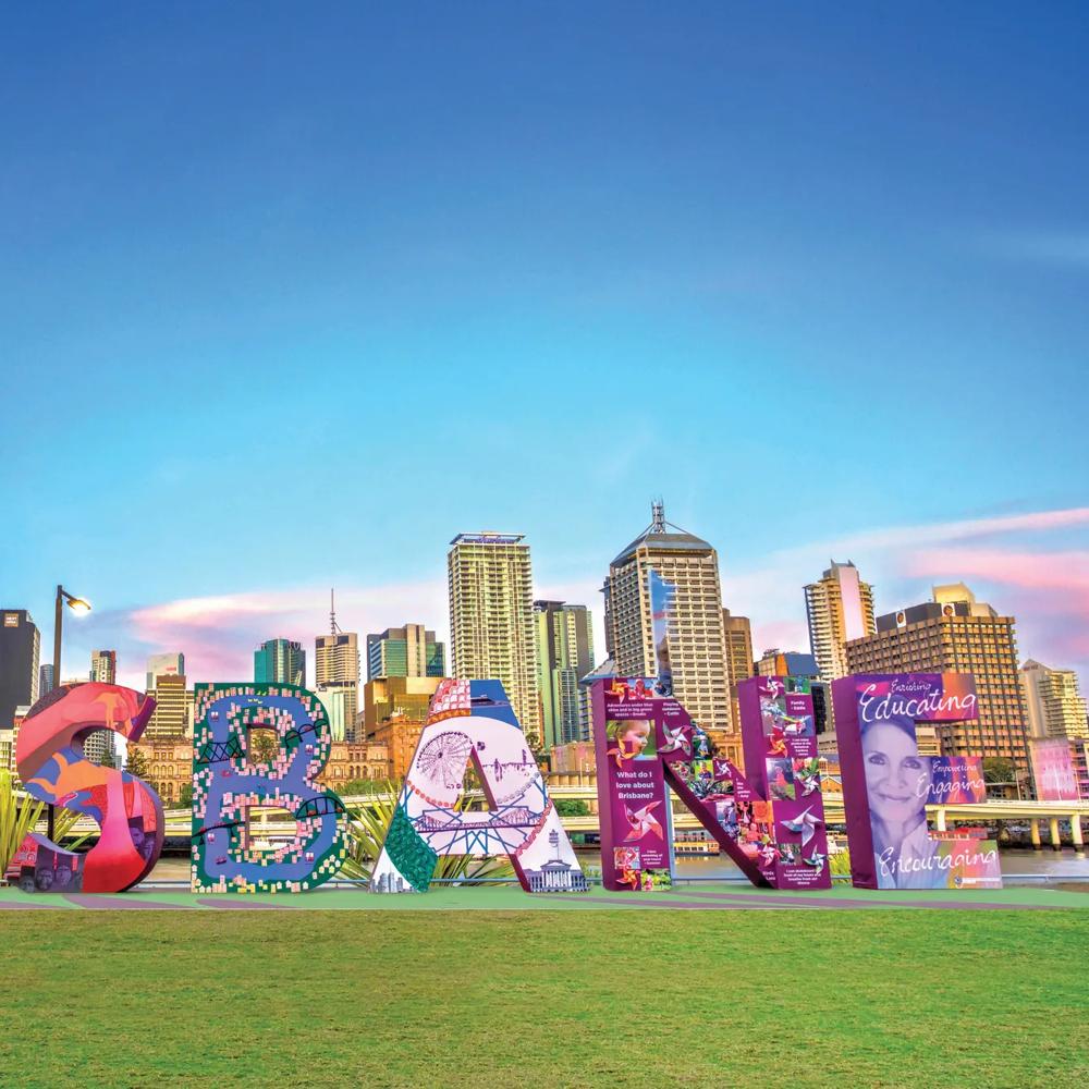 Colourful “BRISBANE” sculpture on a green lawn in front of a skyline with a clear sky.