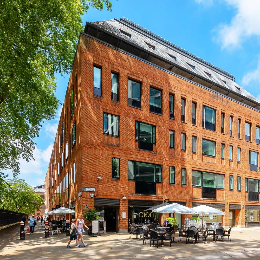 Rotes Backsteingebäude mit großen Fenstern, davor Café mit weißen Sonnenschirmen, umgeben von Bäumen.