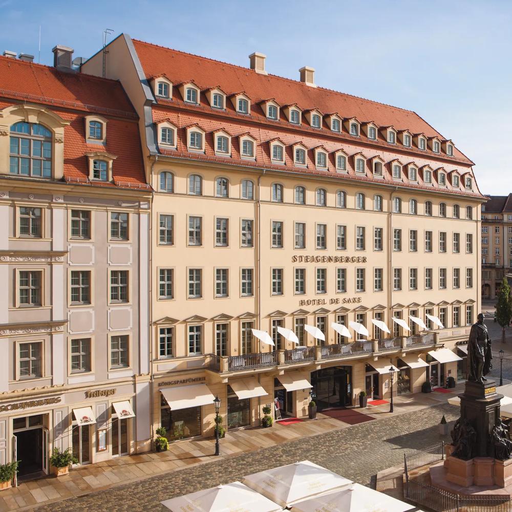 Historischer Platz mit Statue, umgeben von Gebäuden mit roten Dächern und Straßencafés mit weißen Sonnenschirmen.