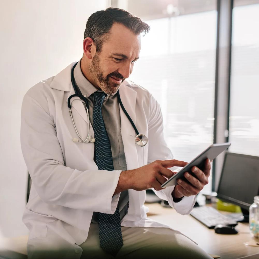 Ein Arzt im weißen Kittel benutzt ein Tablet in einem Büro neben einem Schreibtisch mit Computer.