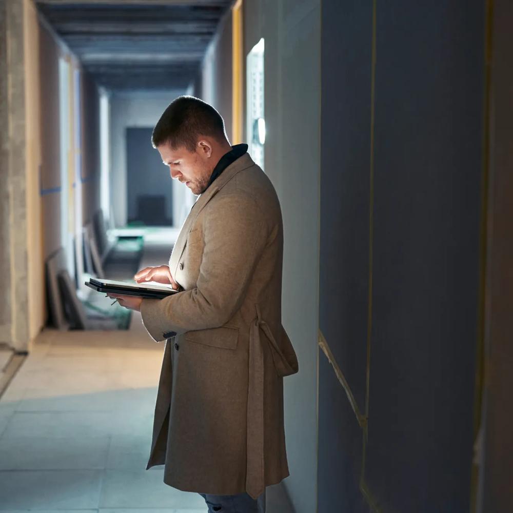 Ein Mann im Mantel steht in einem Flur und tippt auf ein Tablet.