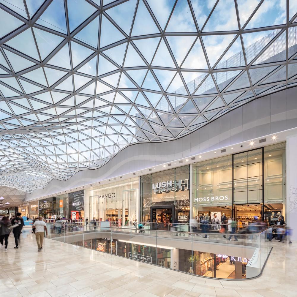 Eine moderne Einkaufspassage mit gläsernem Dach, Geschäften und Menschen, die entlang eines Geländers schlendern.