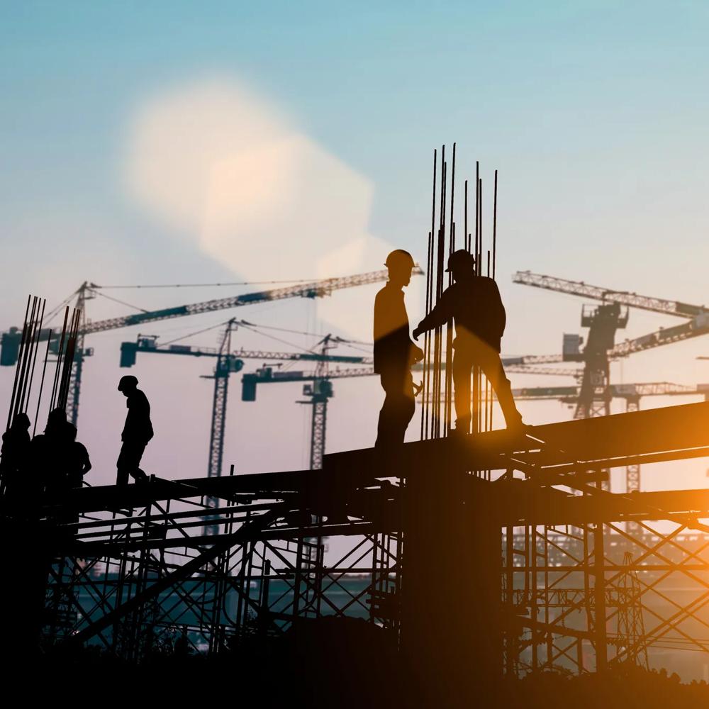 Bauarbeiter auf Gerüst bei Sonnenuntergang, Kräne im Hintergrund.