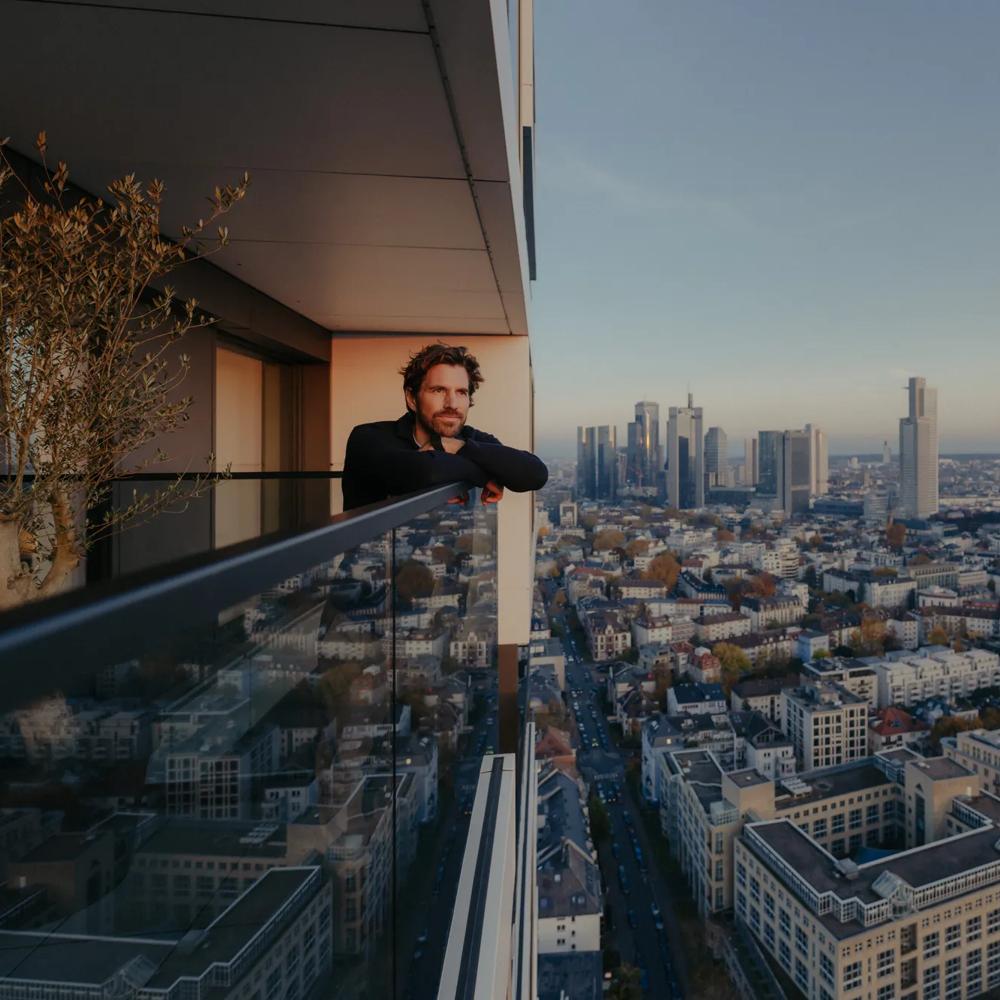Person auf Balkon einer modernen Wohnung mit Blick über die Stadt und Getränk in der Hand.