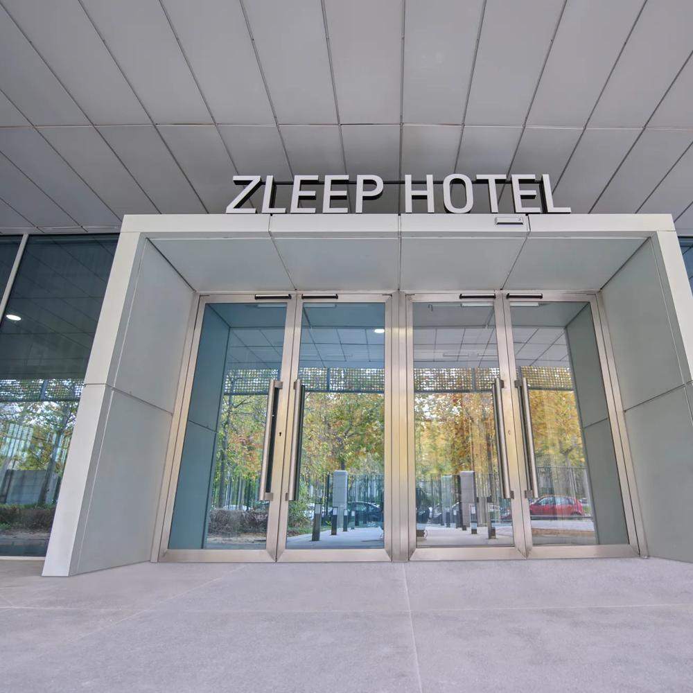 Eingang des Zleep Hotels mit Glasdrehtüren und reflektierenden Fenstern, Bäume im Hintergrund.