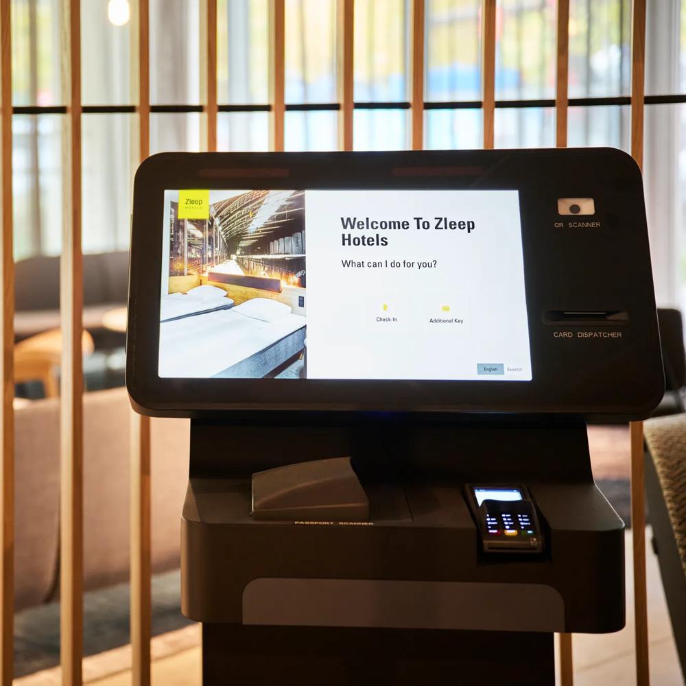 Ein Check-in-Automat in einem Hotel mit Bildschirmtext 'Welcome to Zleep Hotels' vor Holzlatten.