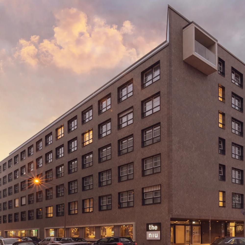 Modernes Gebäude mit vielen Fenstern bei Sonnenuntergang; beleuchtetes Schild und bunte Neonlichter an der Fassade.