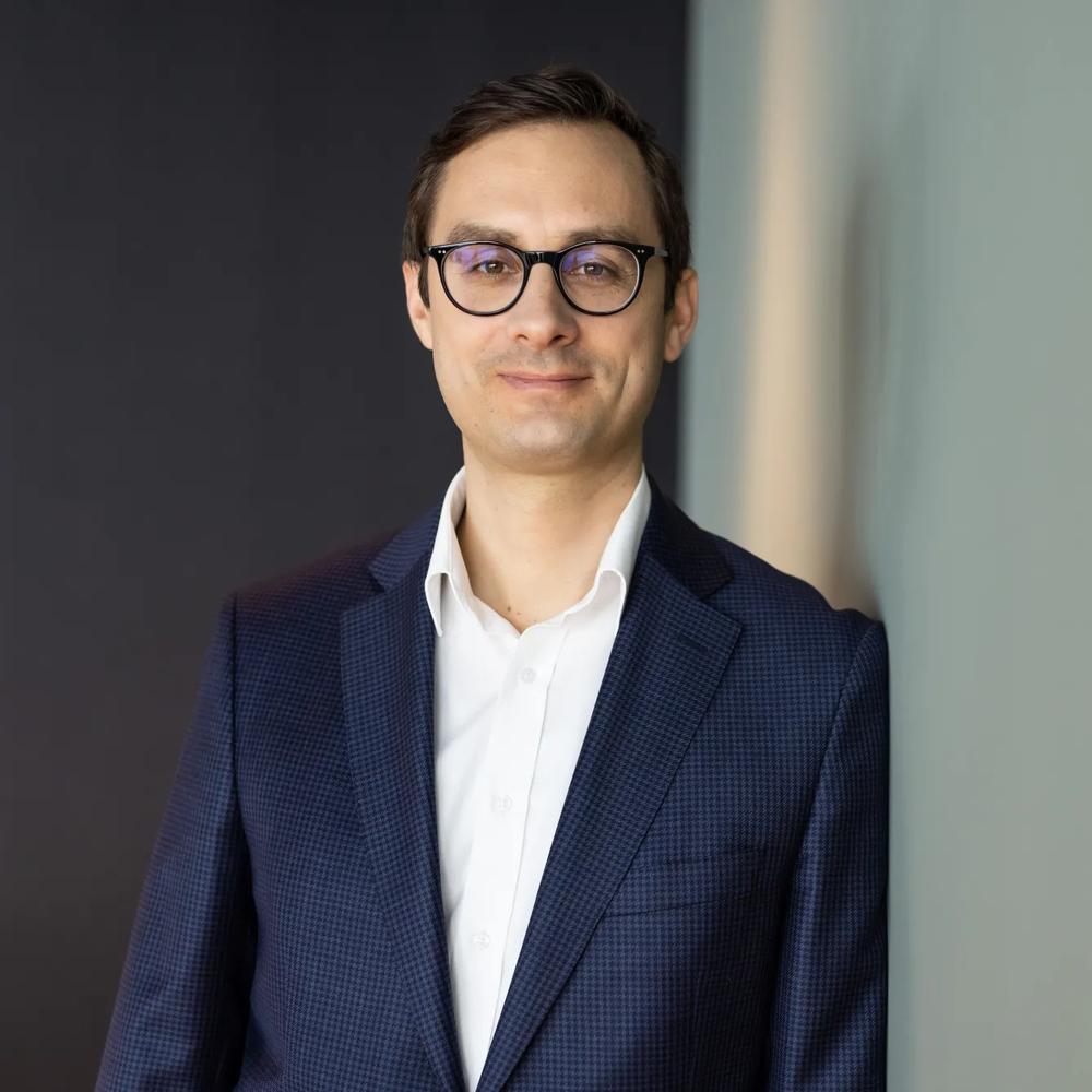 Ein Mann mit Brille im blauen Anzug steht lächelnd vor einer grauen Wand.