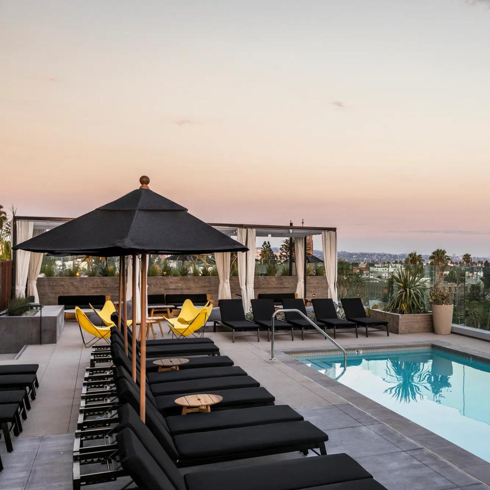 Dachterrasse mit Pool, schwarzen Liegestühlen, gelben Stühlen und Blick auf die Stadt bei Sonnenuntergang.