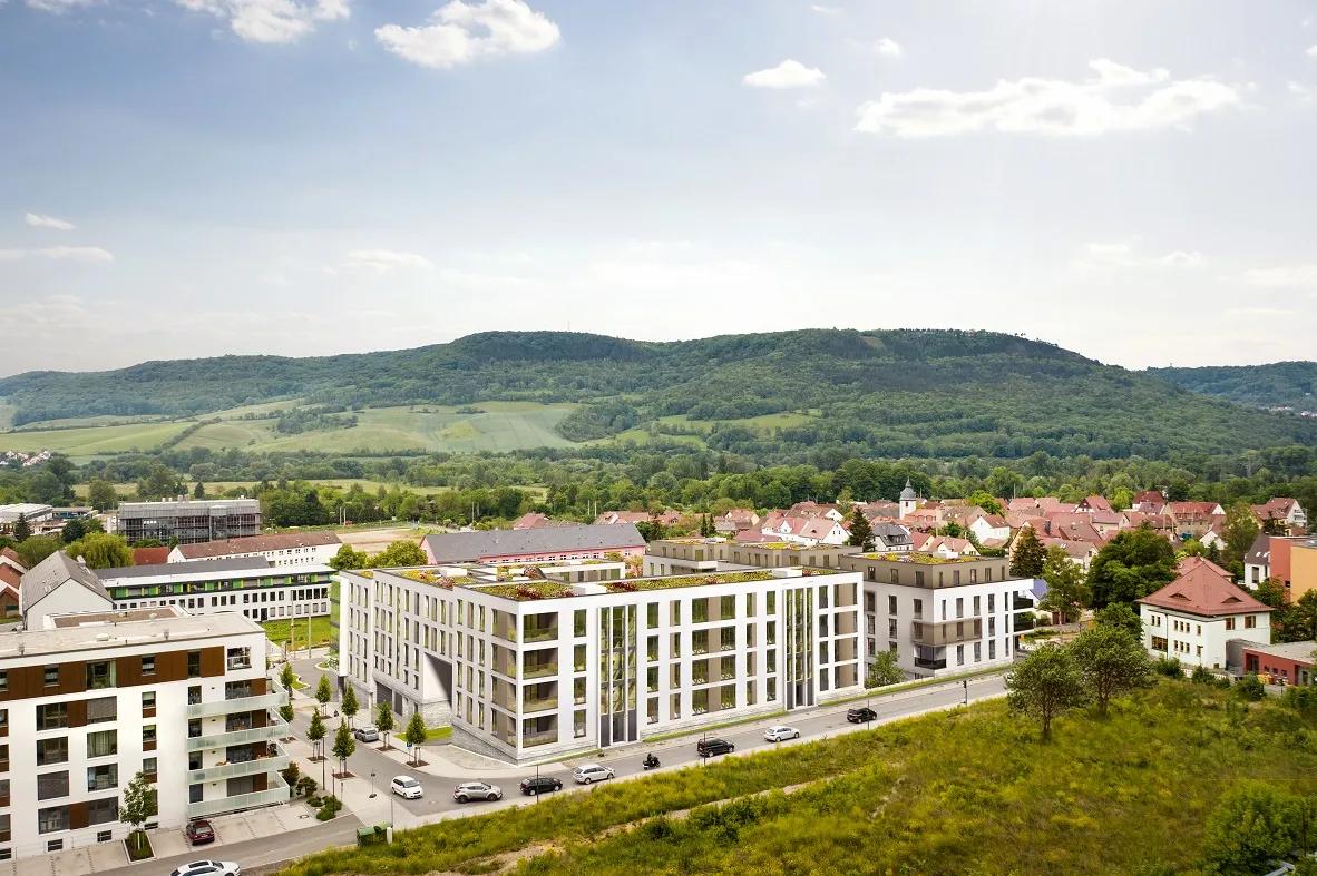 Modern buildings in front of green hills and a village with red roofs under a cloudy sky.