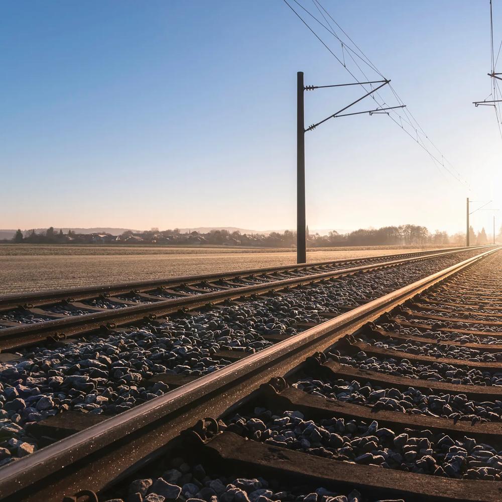 Gleise führen in den Sonnenaufgang, flankiert von Strommasten auf einem frostigen Feld.
