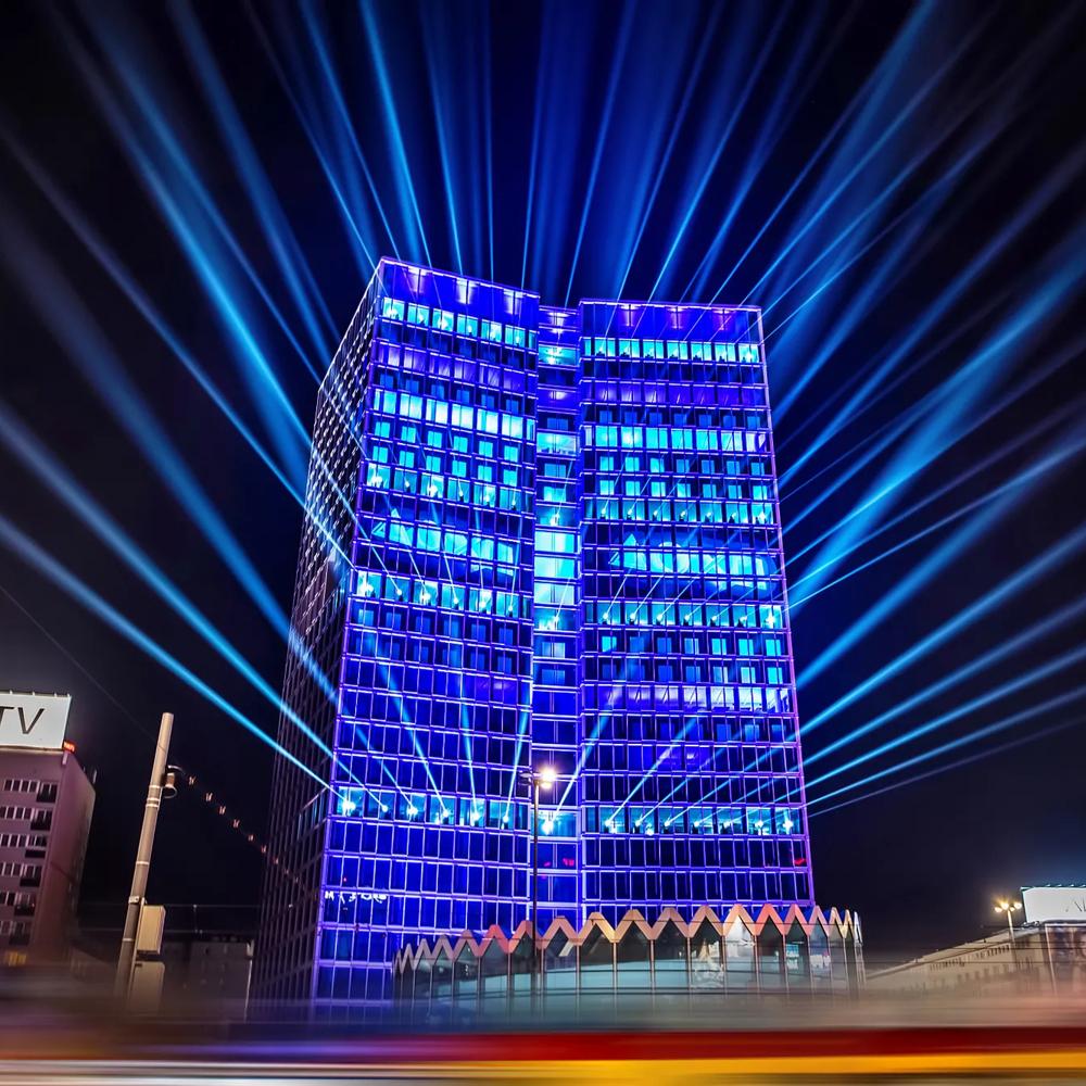 Ein beleuchtetes Hochhaus mit blauen Lichtern und Strahlen am Nachthimmel, flankiert von Gebäuden.