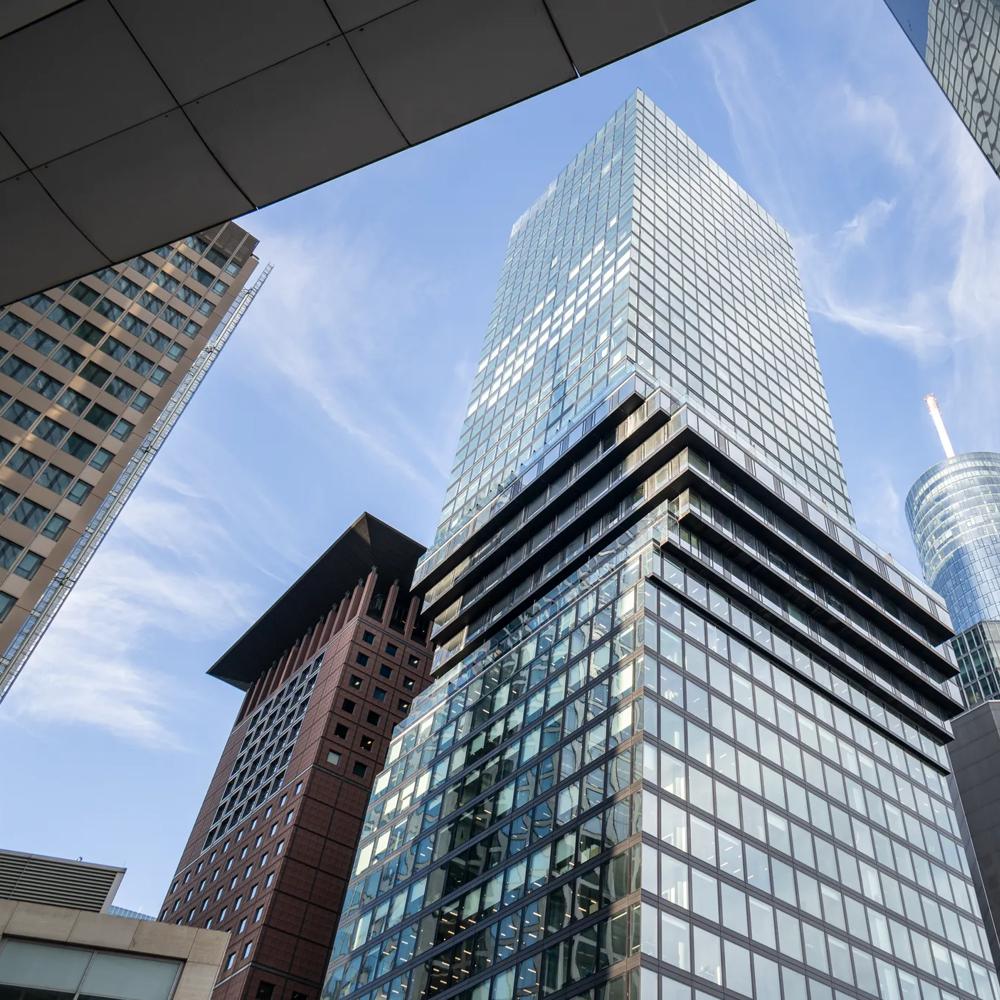 Modernes Hochhaus mit Glasfassade und versetzten Stockwerken zwischen weiteren Hochhäusern.