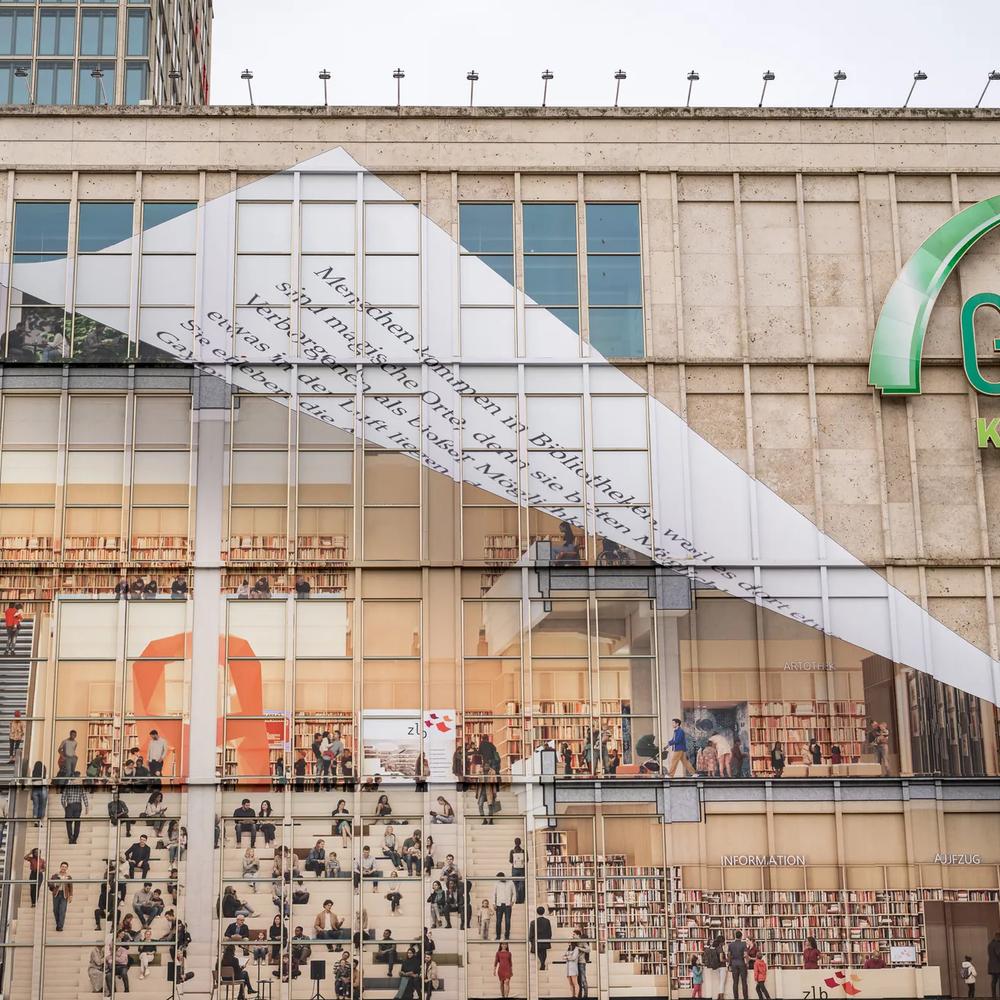 Am Berliner Alexanderplatz wird die Fassade der Galeria-Filiale gezeigt, wie sie von Fassadenkletterern mit bedruckten Klebebahnen versehen wird. die Bahnen visualisieren die mögliche neue Innengestaltung des Gebäudes als offene Bibliothek.