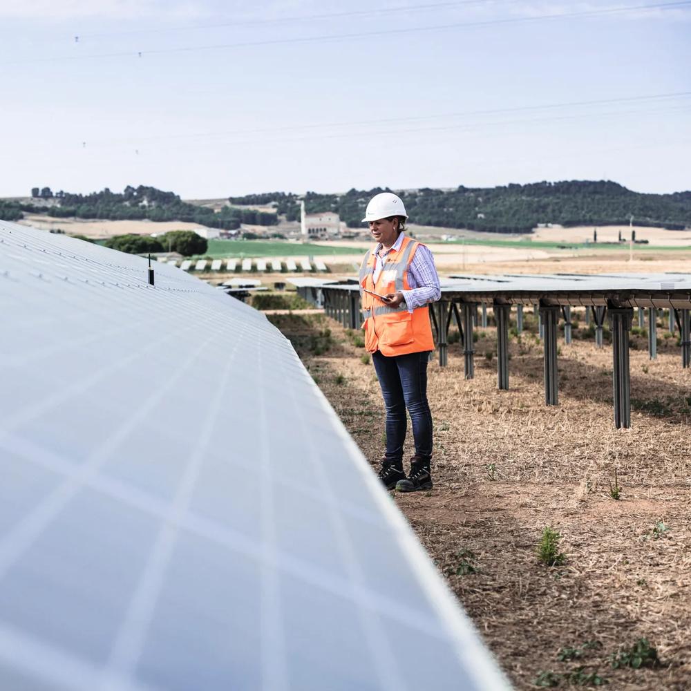 A person wearing protective clothing inspecting solar panels in a field with hills in the background.