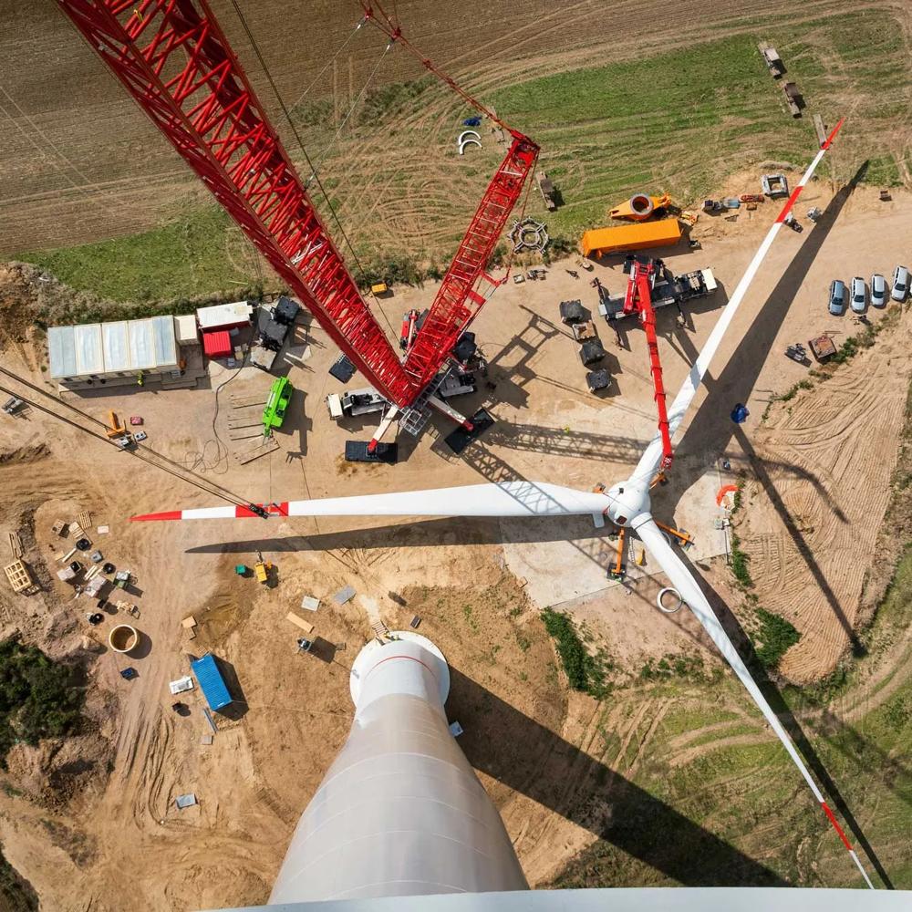 Blick von oben auf eine Windkraftanlage im Bau, mit rotem Kran und Fahrzeugen auf einem Feld.