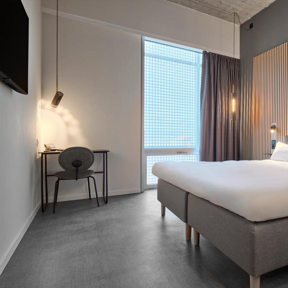 Modernes Hotelzimmer mit großem Bett, Holzpaneelwand, Schreibtisch, Stuhl und hängender Lampe neben einem Fenster.