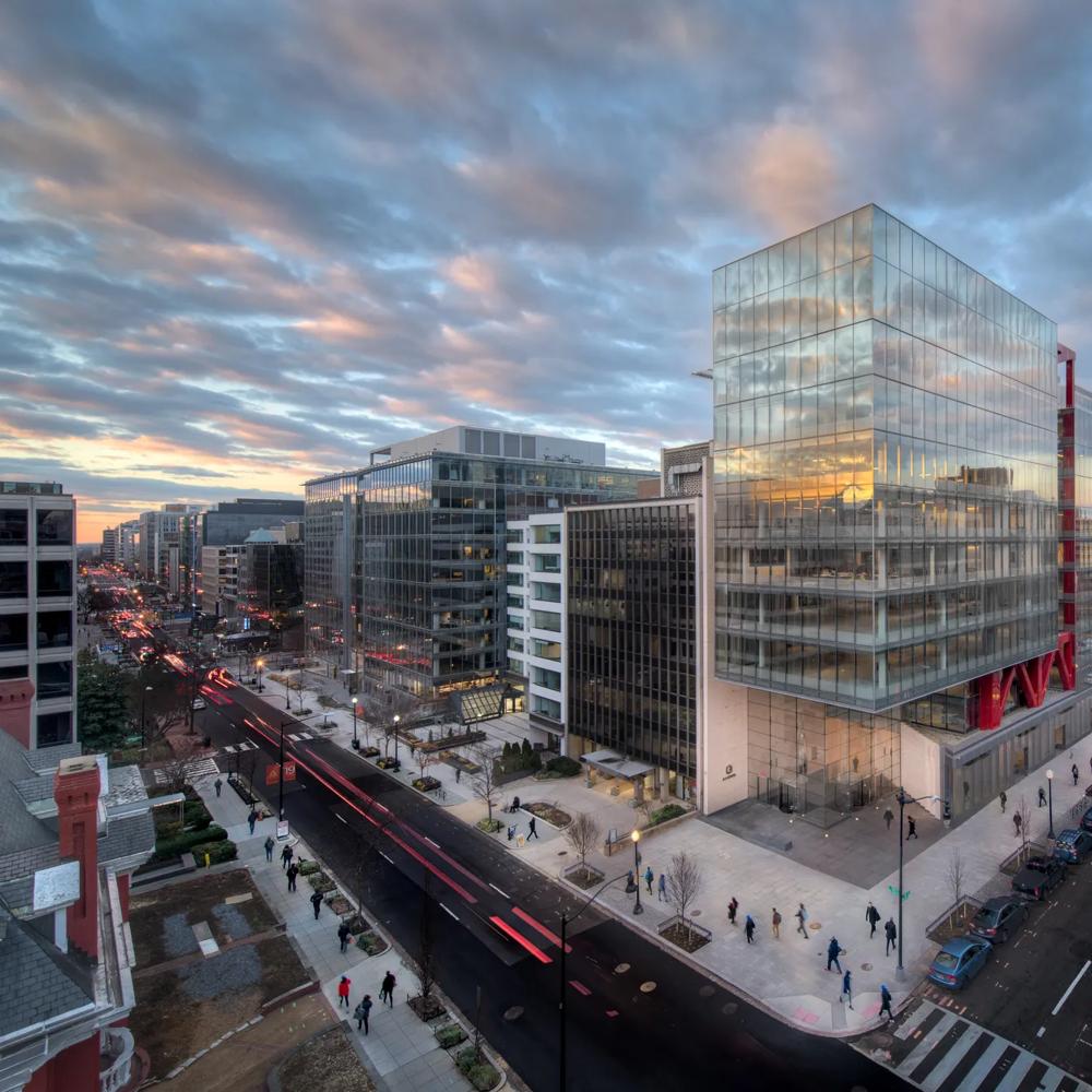 Moderne Glasgebäude bei Sonnenuntergang, Menschen auf Gehwegen, belebte Straßen mit Autos, bewölkter Himmel.