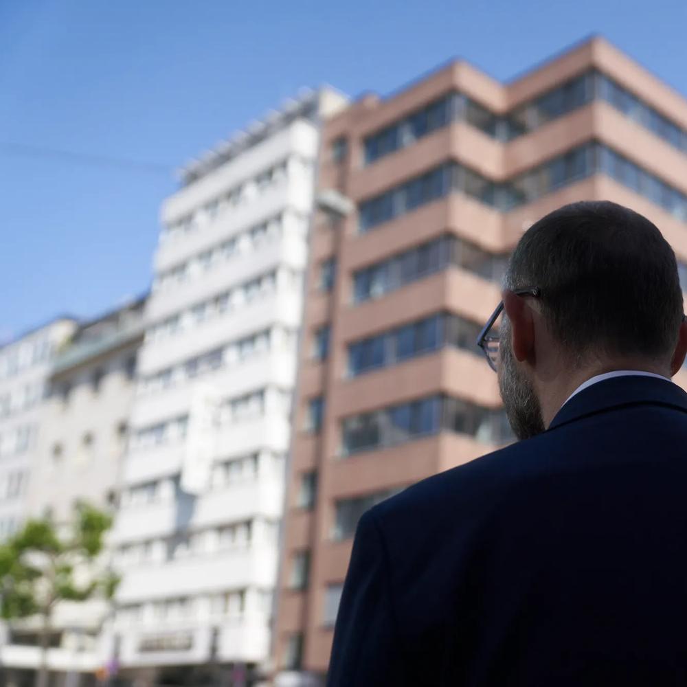Ein Mann im Anzug steht vor modernen Hochhäusern, blauer Himmel im Hintergrund.