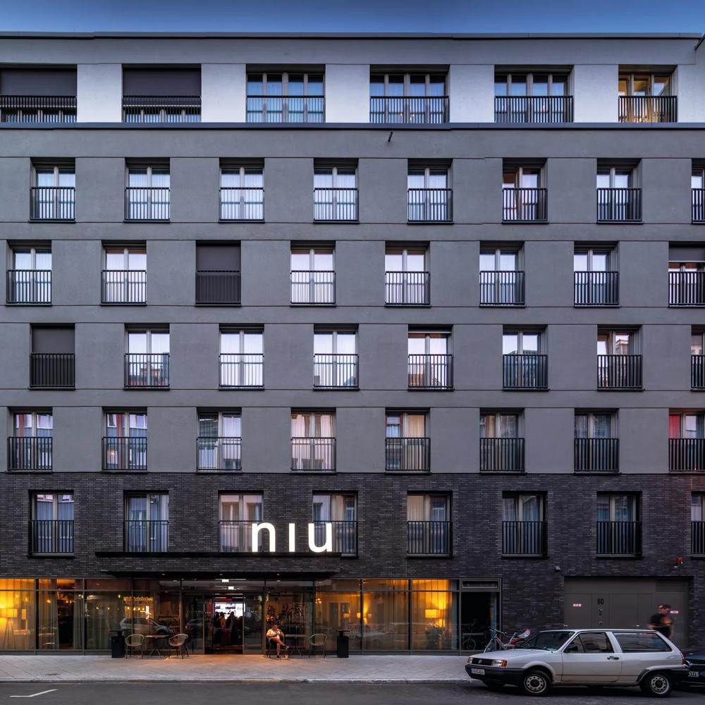 Graues Gebäude mit Balkonen, Leuchtreklame "niu" und rotem Schild, Autos parken davor.