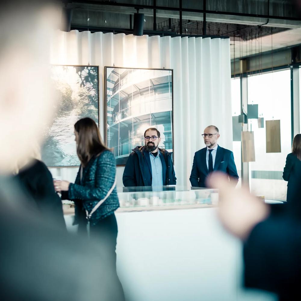 Networking-Szene in einem modernen Ausstellungsraum mit Architekturfotos und einem Gebäudemodell. Zwei Männer stehen im Zentrum, umgeben von Gästen in Business-Kleidung.