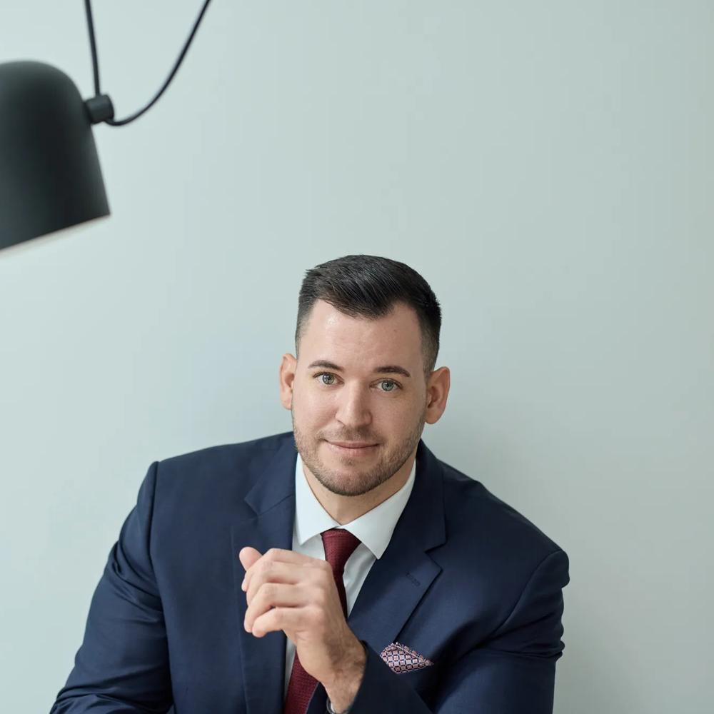 Ein Mann trägt einen schwarzen Anzug mit einer roten Krawatte und einem Einstecktuch. Er sitzt vor einer mintfarbenen Wand, neben ihm ragt eine schwarze Lampe ins Bild.