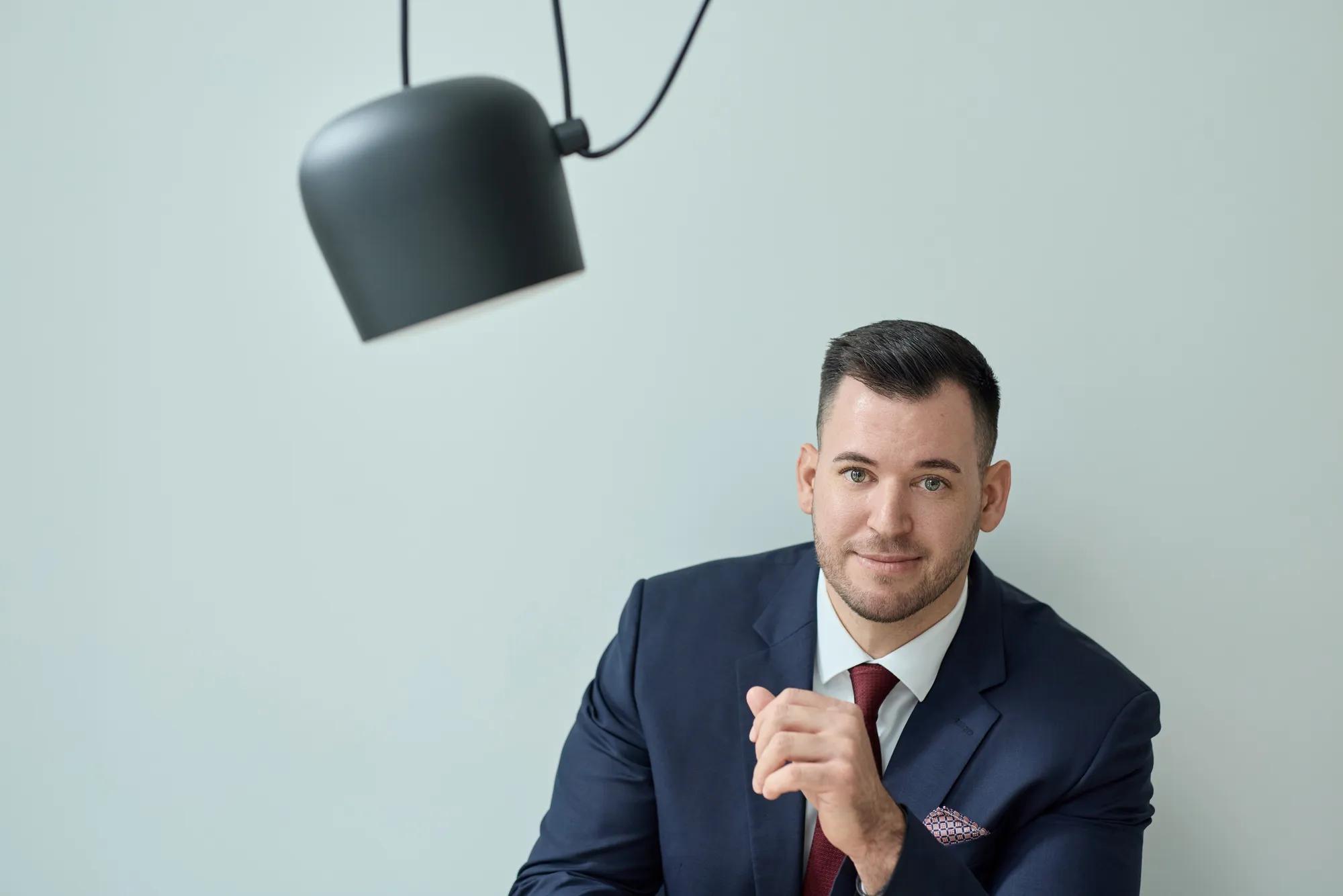 Ein Mann trägt einen schwarzen Anzug mit einer roten Krawatte und einem Einstecktuch. Er sitzt vor einer mintfarbenen Wand, neben ihm ragt eine schwarze Lampe ins Bild.