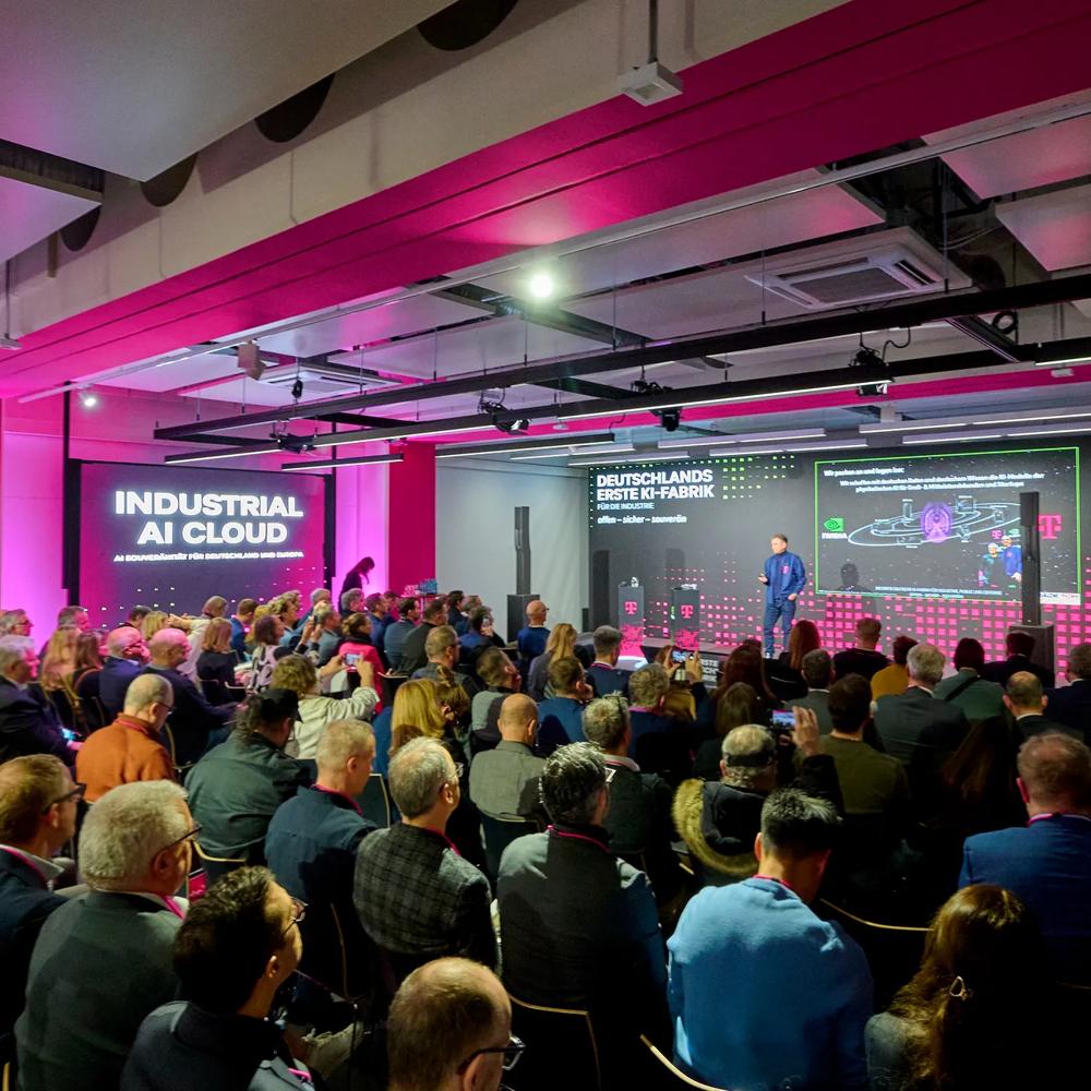 Vollbesetzter Veranstaltungssaal in magentafarbenem Licht bei der Vorstellung der „Industrial AI Cloud" – einer Kooperation von Deutsche Telekom und NVIDIA. Ein Redner präsentiert auf der Bühne vor großen Screens.