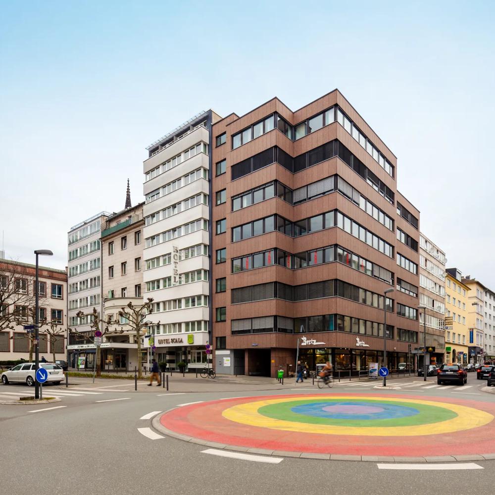 Ein Kreisverkehr mit Regenbogenfarben, umgeben von modernen Gebäuden und Geschäften in einer Stadtstraße.