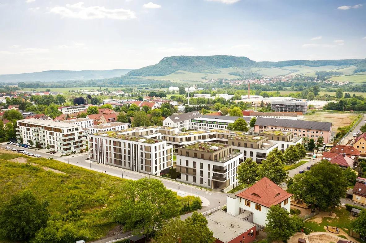 Modern residential building with green roofs in front of a hilly landscape and a small village in the background.
